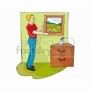 A man in a red shirt and blue pants is straightening a picture on the wall in a home setting. A small table with a drawer and a vase with a flower is beside him.