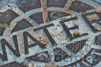 Close-up photograph of a metal utility cover with the word 'WATER' engraved on it. The cover features a weathered texture with small pieces of debris and signs of rust.