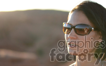 A woman wearing sunglasses with a reflection of a photographer in them, set against a blurry outdoor background with hills.