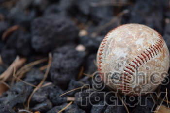 A close-up image of a worn baseball with red stitching resting on dark, rocky ground.