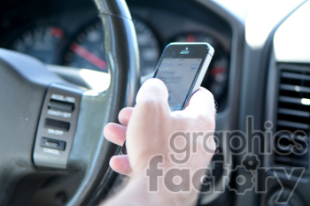 A close-up image of a person using a smartphone while driving. The person's hand is holding the phone with the car's steering wheel and dashboard visible in the background.
