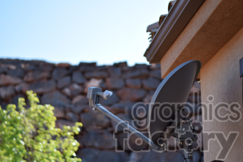 A satellite dish mounted on the side of a house, with a stone wall and greenery in the background under a clear blue sky.