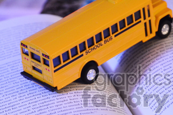 A miniature yellow school bus toy model is placed on an open book, visible in this clipart image. The school bus is detailed with an 'Emergency Exit' sign at the rear.