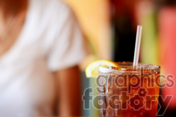 A close-up image of an iced tea with a lemon wedge and a straw, with a blurred background featuring a person in a white shirt.