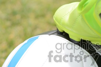 Close-up image of a soccer ball with blue and black stripes, with a neon green soccer cleat resting on top. The background features blurred grass, indicating an outdoor setting.