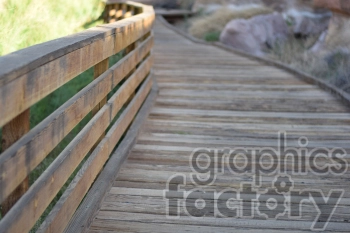 A wooden boardwalk with a railing curves gently through a natural outdoor setting with greenery and rocks.