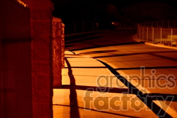 A nighttime view of a dimly lit sidewalk and street with shadows cast by fencing and brick pillars, illuminated by a warm orange streetlight.