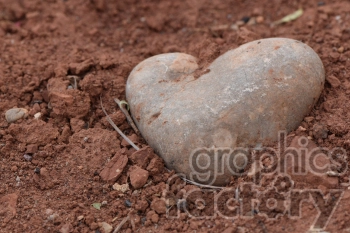 A heart-shaped stone partially buried in reddish soil and surrounded by small rocks and dirt clods.
