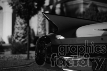 A black and white image of the rear part of a classic car with prominent tailfins. The car is parked outdoors, and the photo is taken at night, highlighting the distinct design of the vehicle.