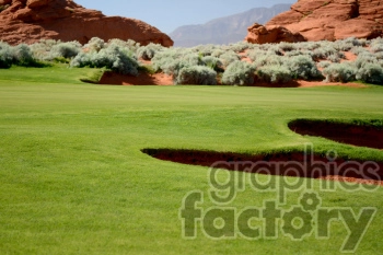 A scenic view of a golf course with a bunker surrounded by green grass and desert vegetation, set against a backdrop of red rock formations and distant mountains.