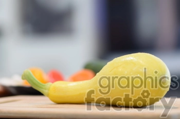 A fresh yellow squash resting on a wooden cutting board, with blurred vegetables in the background.