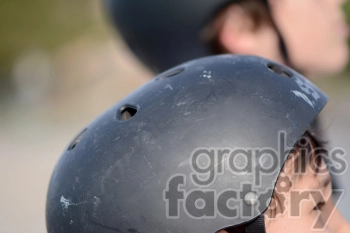 Close-up image of two people wearing black helmets. The focus is on the texture and details of the helmet in the foreground, with another helmet visible in the blurred background.
